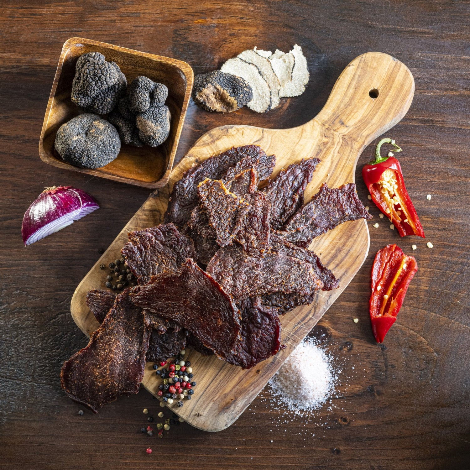 Matt-Hat Jerky's Black Truffle Beef Jerky on a wooden slab with black truffle, onions, peppers, and spices around it.