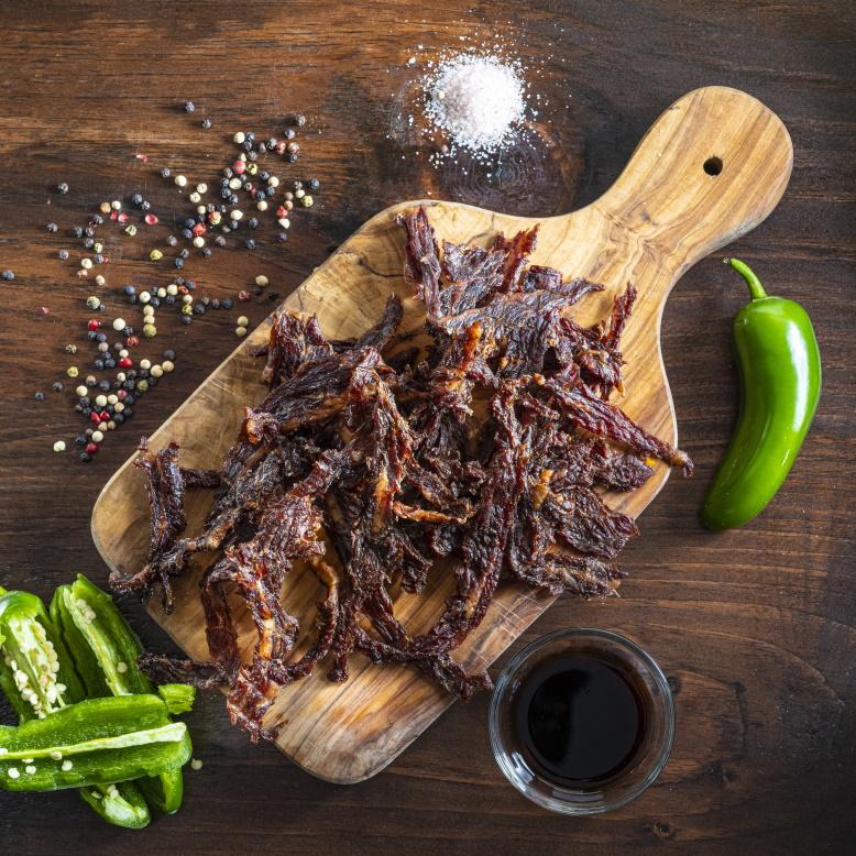 Matt-Hat Jerky's Gourmet Keto Original Beef Jerky on a wooden slab with peppers, sauces, and spices around it.